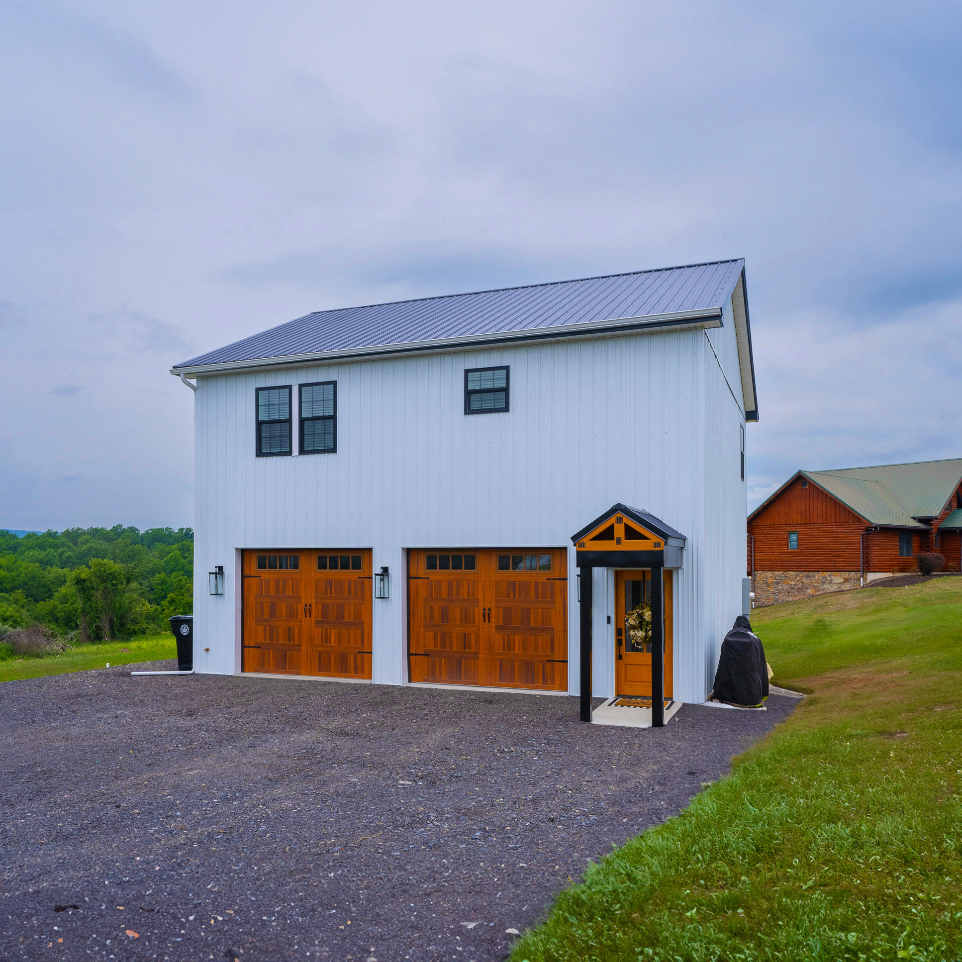 Two-Story Barndominium with Living Quarters Over Garage – PA White barndominium with upper-level living space and wood-style garage doors, built by a Mid-Atlantic pole barn company in Pennsylvania